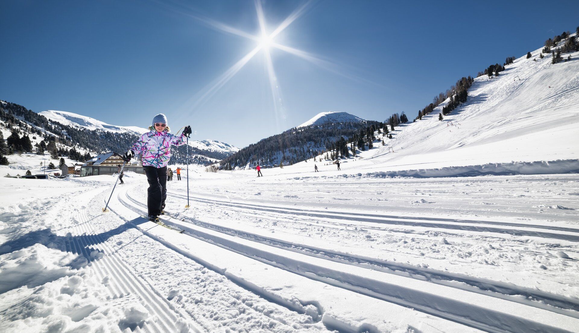 Ein Langläufer auf einem verschneiten Pfad, mit blauem Himmel und Bergen im Hintergrund.