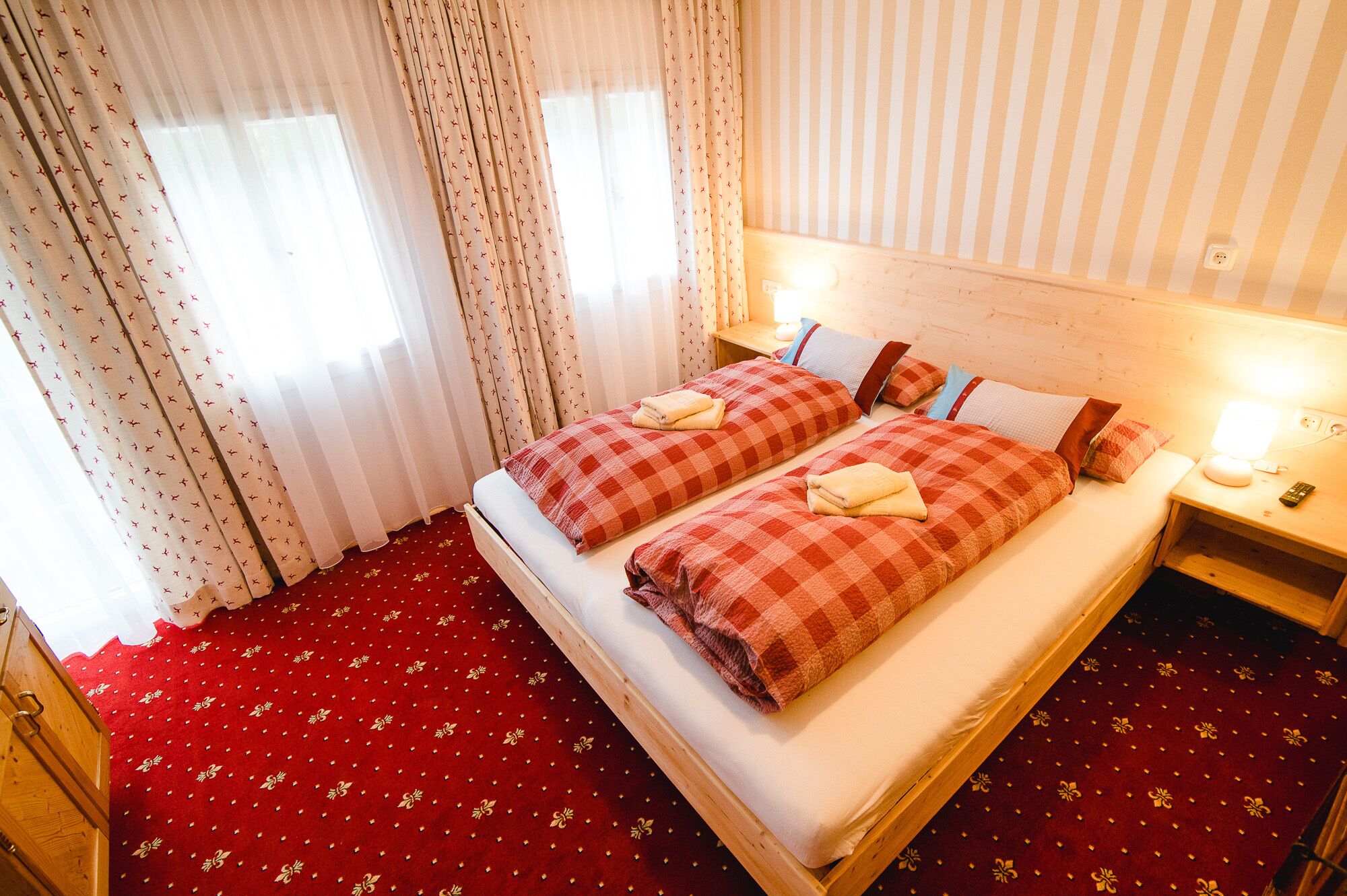 Een tweepersoonsbed met geruit beddengoed, een houten hoofdbord, een nachtkastje met een lamp en een rode vloerbedekking.