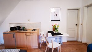 A compact kitchen with a sink, a coffee maker, and a small refrigerator. There's a white table with a vase of flowers and a chair. A door is visible in the background. - Apartment 01 Rauenstein (Rathen)