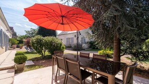 Um pátio com mesa e cadeiras, um guarda-sol vermelho e um jardim com diversas plantas e uma árvore.
