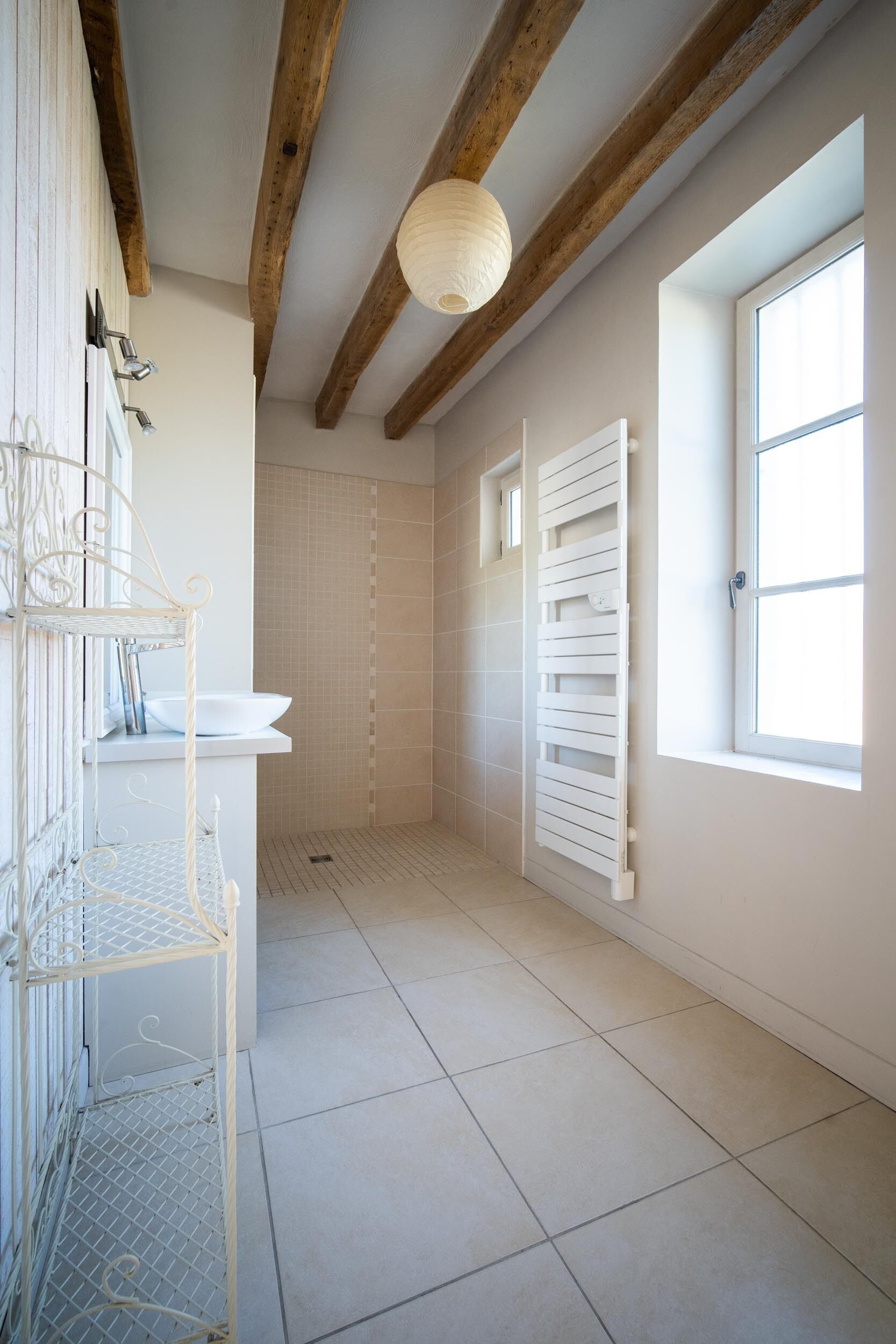 Une salle de bain avec un lavabo blanc, un porte-serviettes blanc, une douche avec une porte blanche et une fenêtre laissant entrer la lumière naturelle.