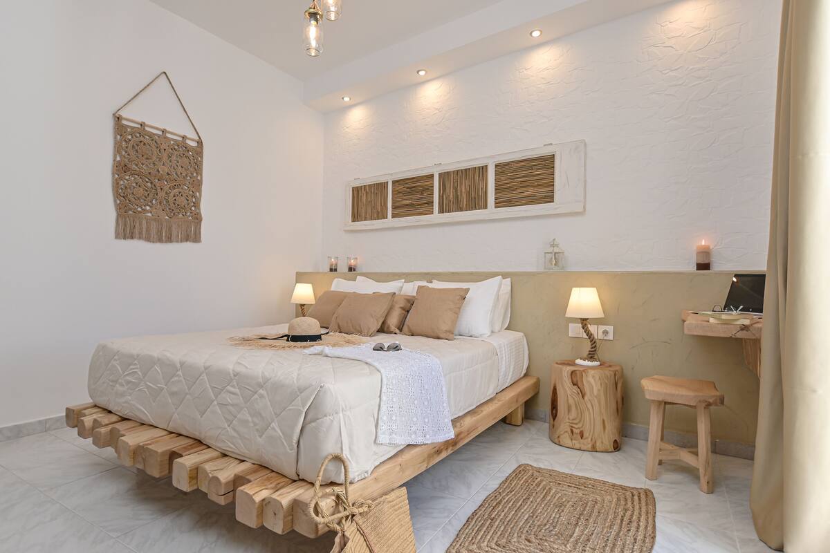 Quarto com estrutura de cama de madeira, mesa de cabeceira com abajur, um banquinho pequeno e um enfeite na parede.