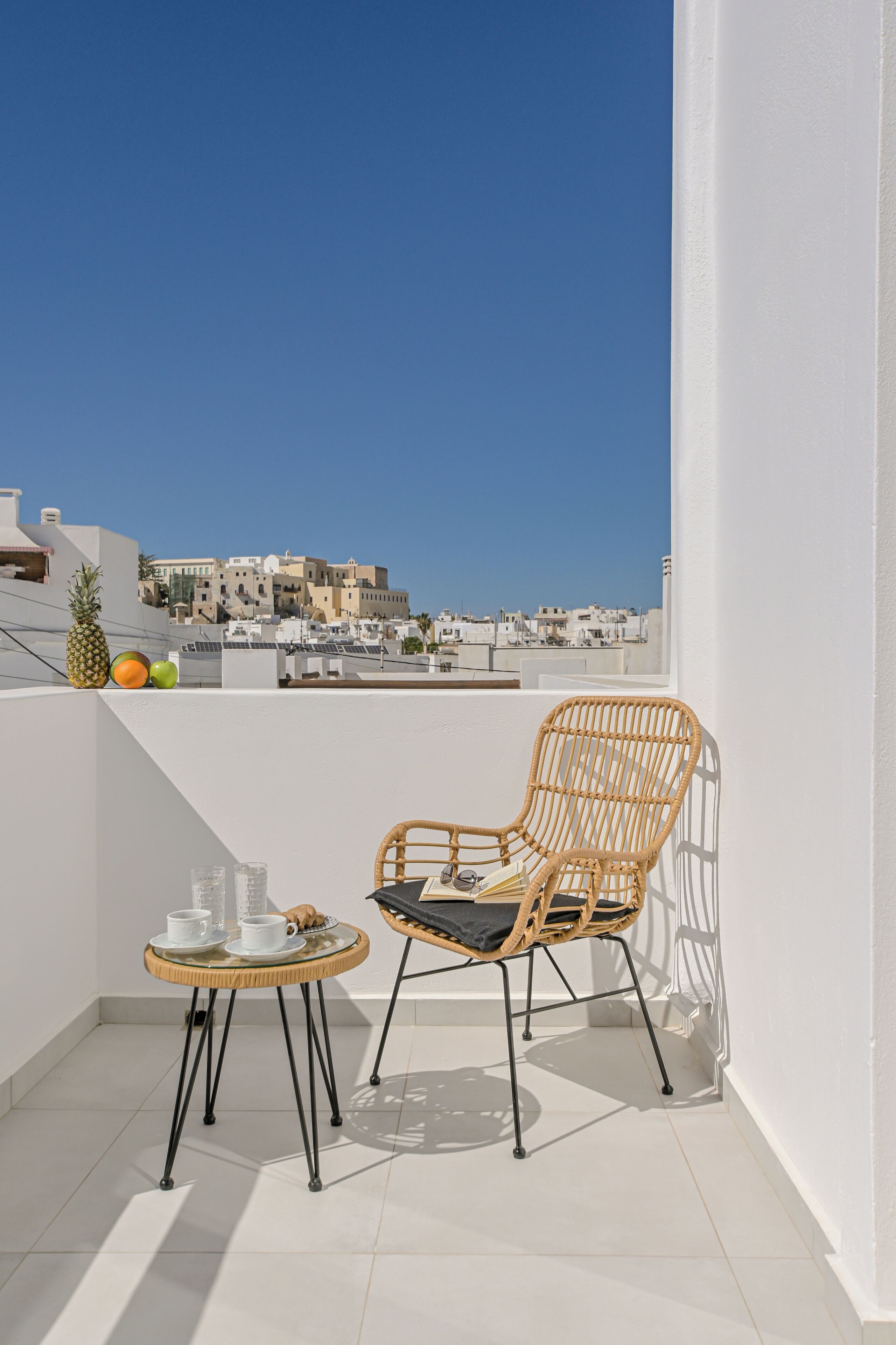 Un balcon avec un fauteuil en osier, une petite table ronde avec une tasse et une soucoupe blanches, et un ananas.