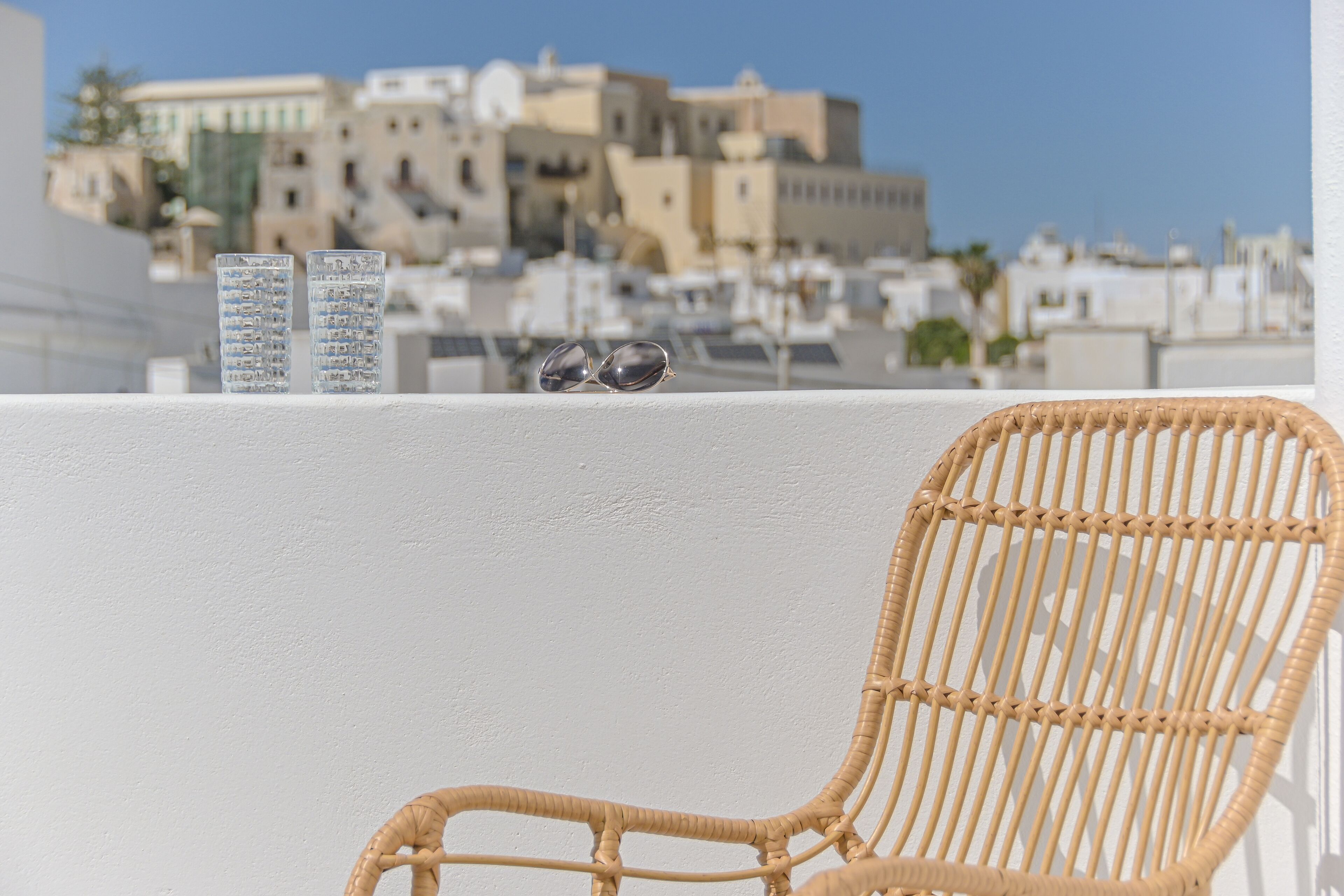 Une chaise en osier et deux verres sur un balcon, avec en arrière-plan une vue sur des immeubles.
