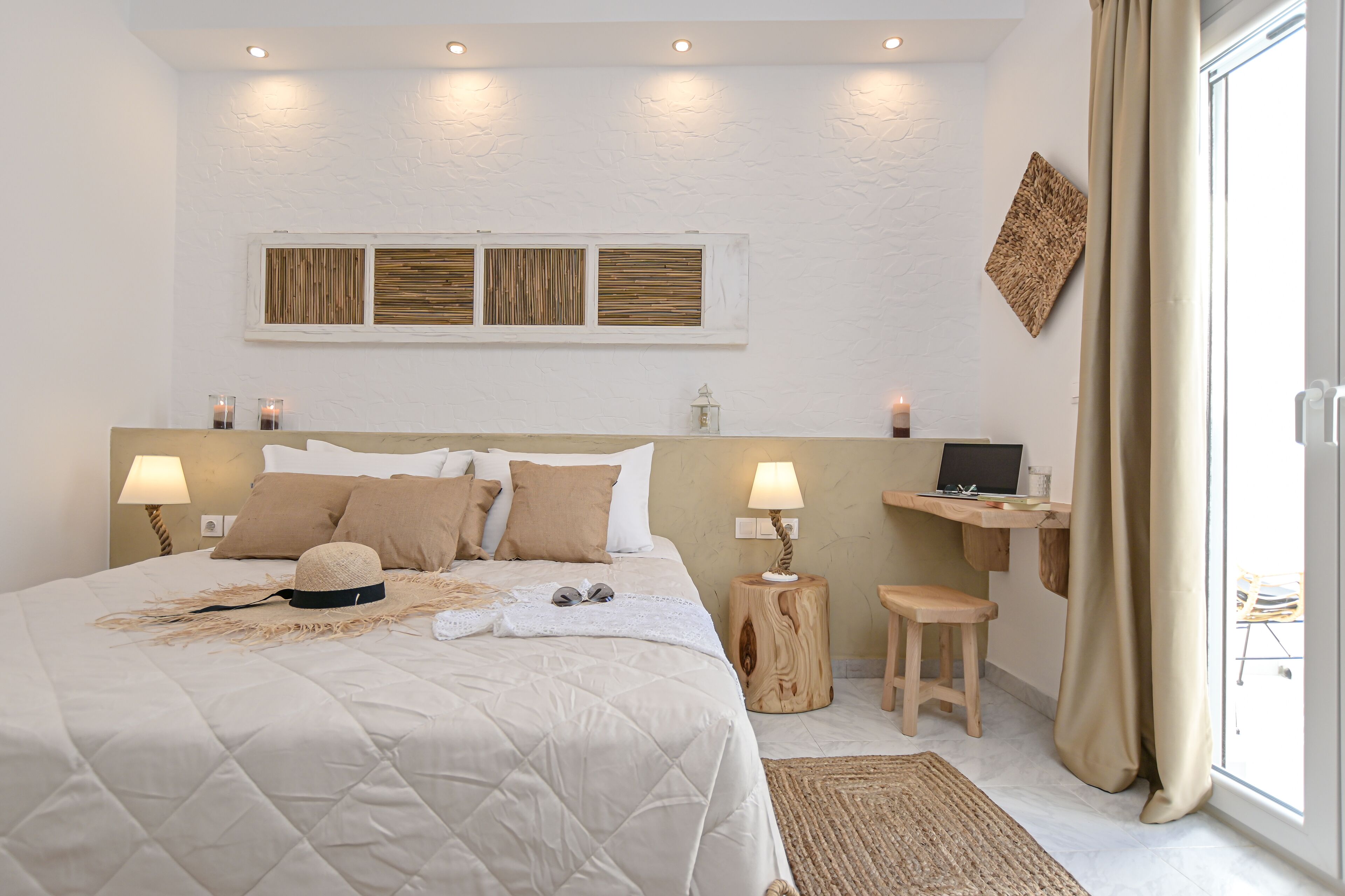 A bedroom with a large bed, two bedside lamps, a laptop on a wooden side table, and a view through a window with curtains.