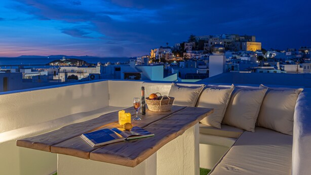 A rooftop terrace with a wooden table, a white sofa, and a view of a coastal town at dusk. - Naxian Horizon (Naxos)