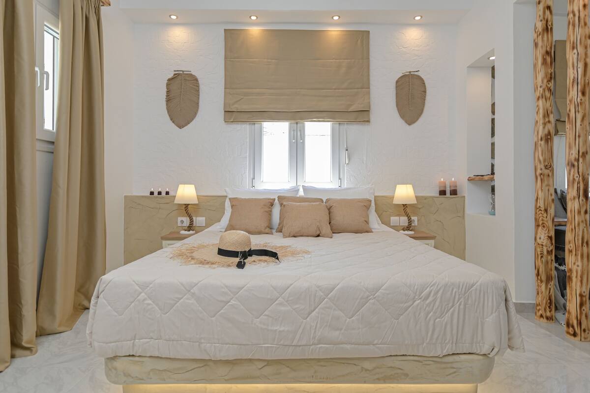 Quarto com uma cama grande, duas mesas de cabeceira, uma janela com persiana bege e uma folha decorativa na parede.