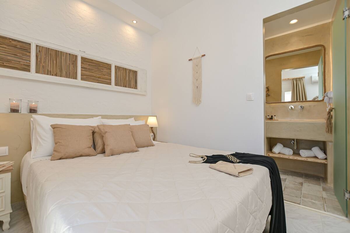 Quarto com uma cama grande, mesas de cabeceira, um enfeite de parede em macramé, um espelho e um banheiro com pia e porta-toalhas.