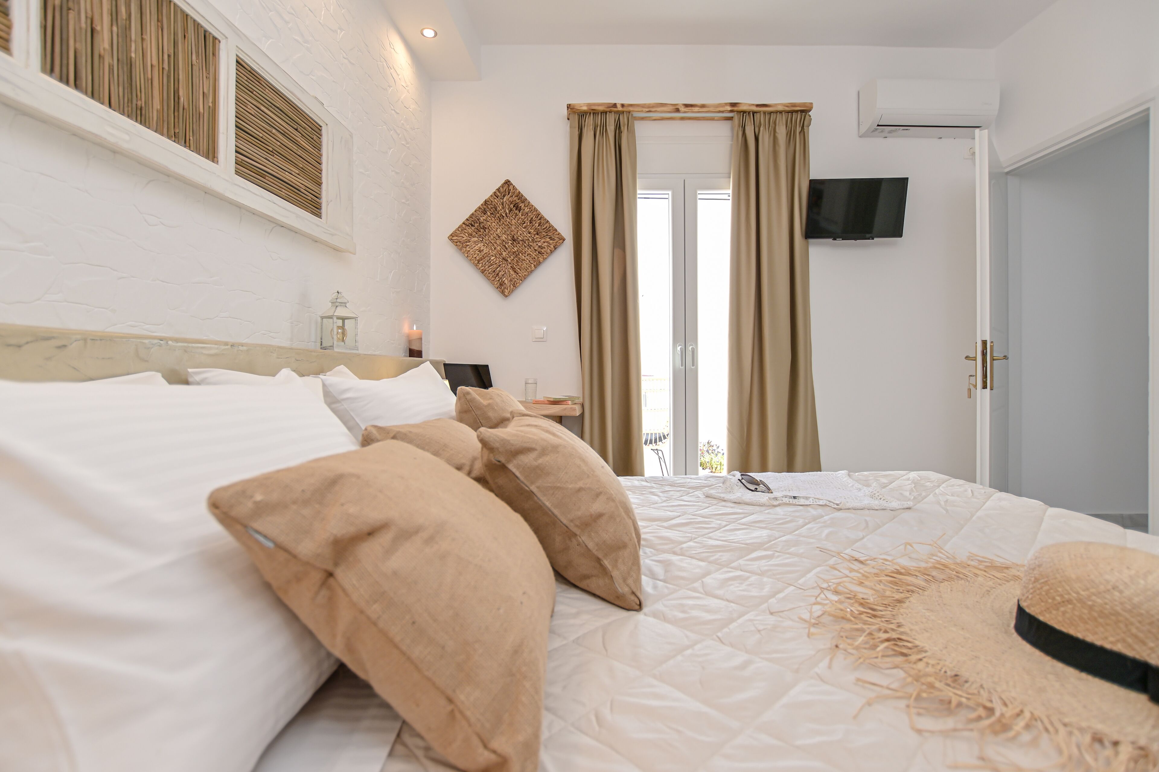 Een netjes opgemaakt bed met beige en witte beddengoed, een aan de muur bevestigde televisie, en een raam met gordijnen.
