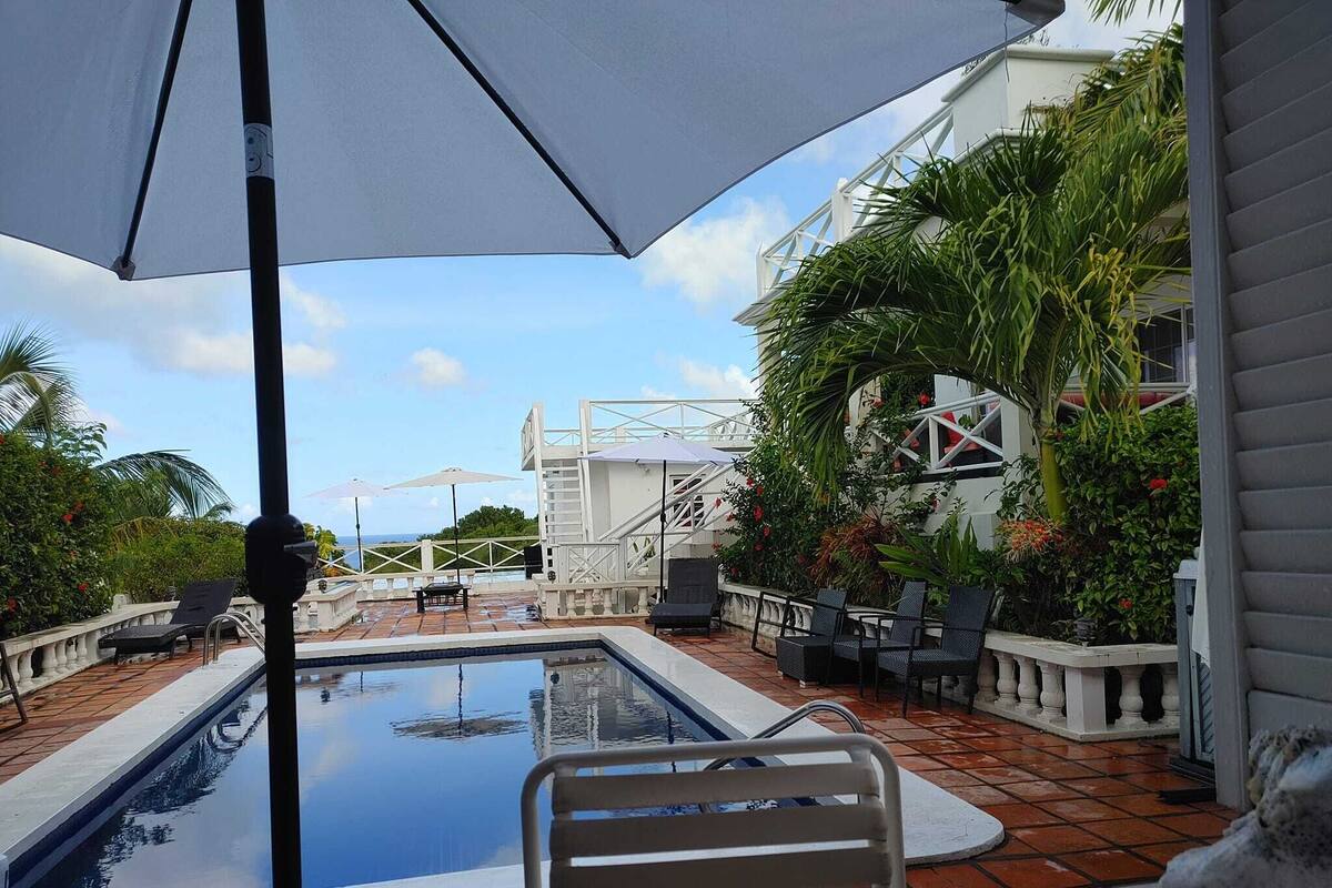 A pool area with a view of the ocean, surrounded by lush greenery and a white building with a staircase.