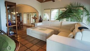 A spacious living room with a large white sectional sofa, a wooden ceiling fan, and a large window with blinds. - Chez Mango Villas (Old Towne)