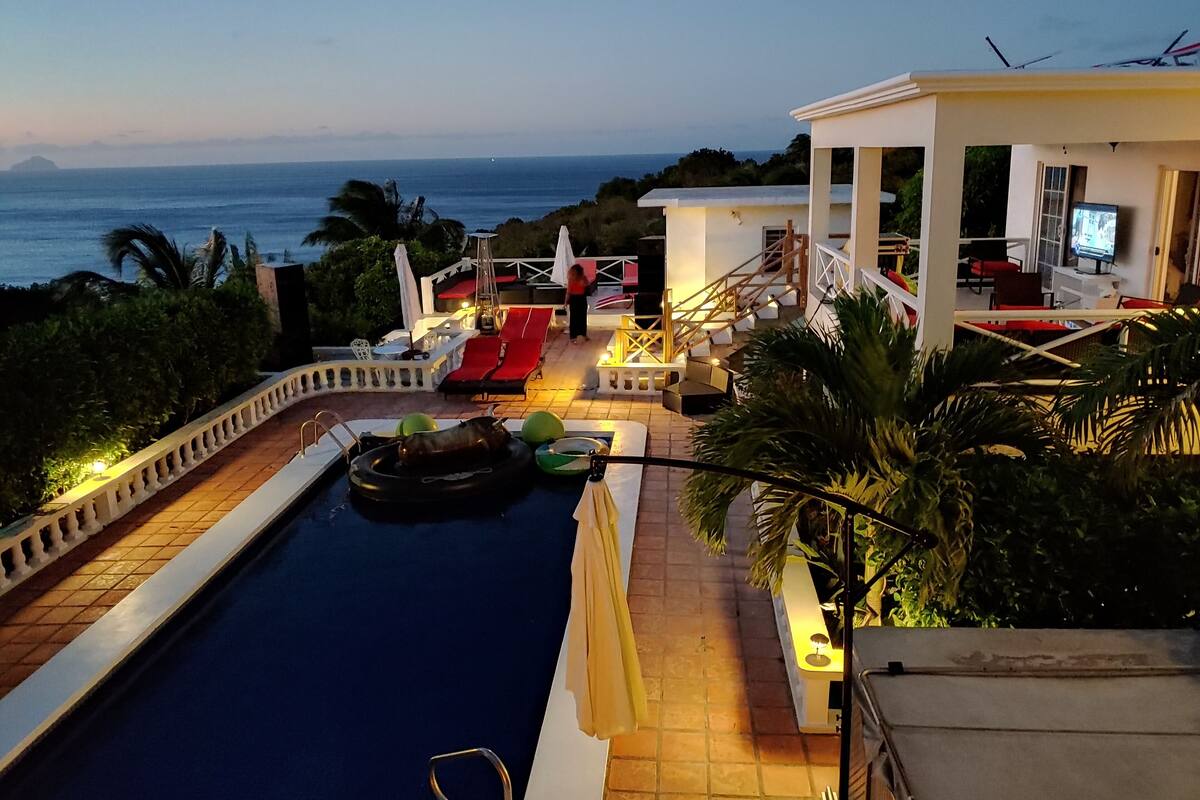 A pool area with a view of the ocean, a lounge with red chairs, and a white building with a staircase.