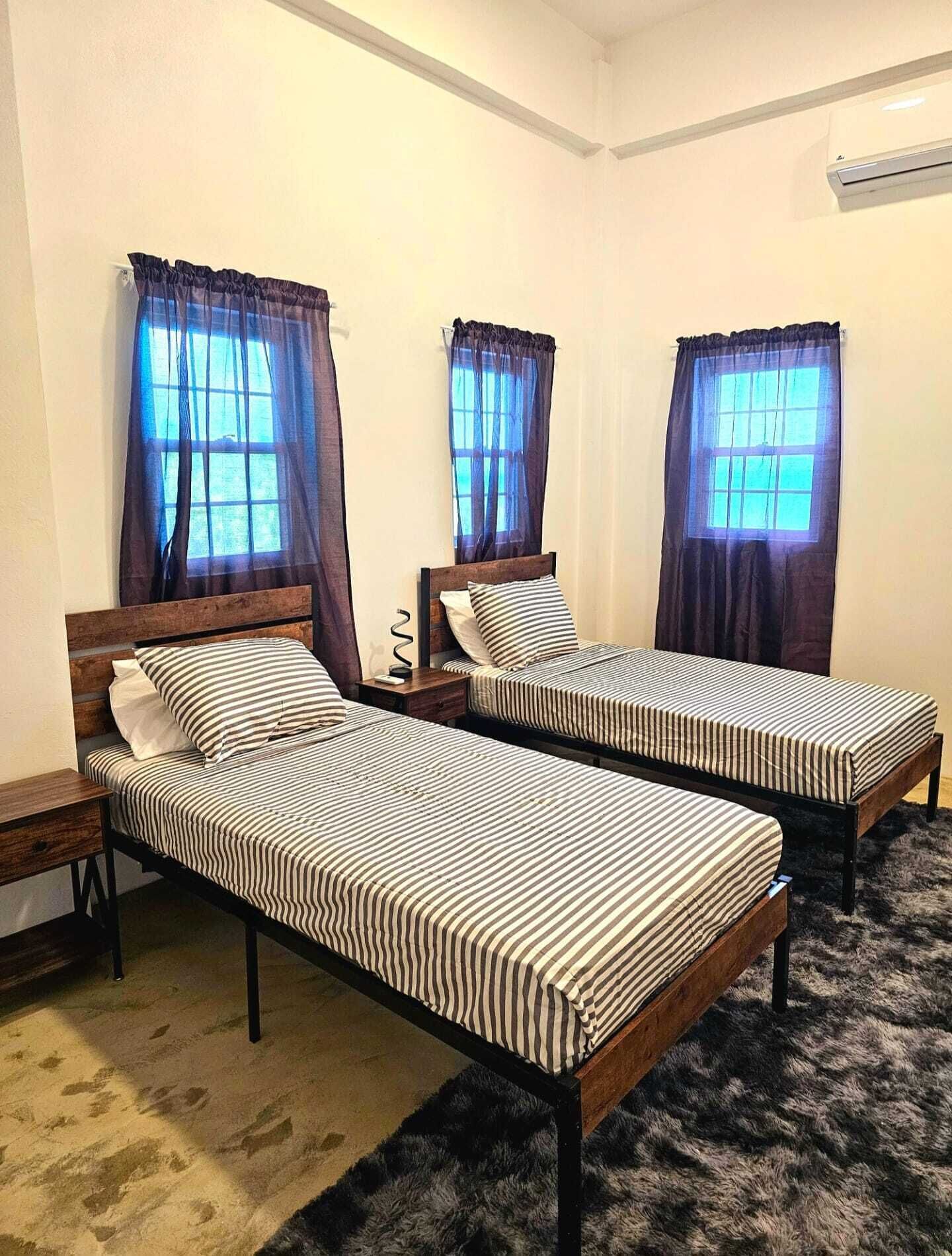 Ein Zimmer mit zwei Einzelbetten, jeweils mit einem hölzernen Kopfteil und gestreifter Bettwäsche. Zwei Fenster mit Vorhängen und ein kleiner Nachttisch mit Lampe.
