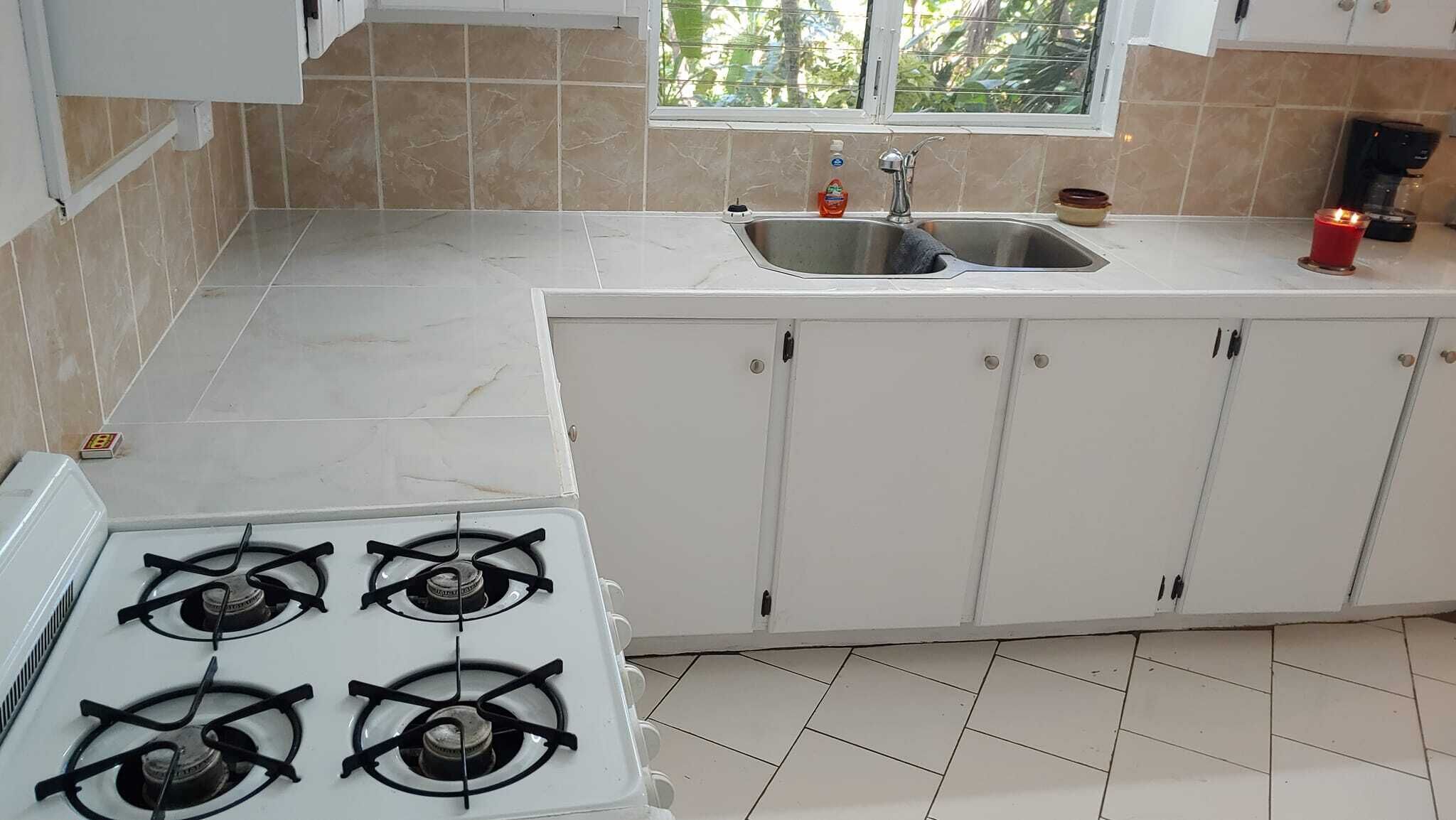 Cocina con alacenas blancas, estufa a gas, lavamanos y ventana con vista a áreas verdes.