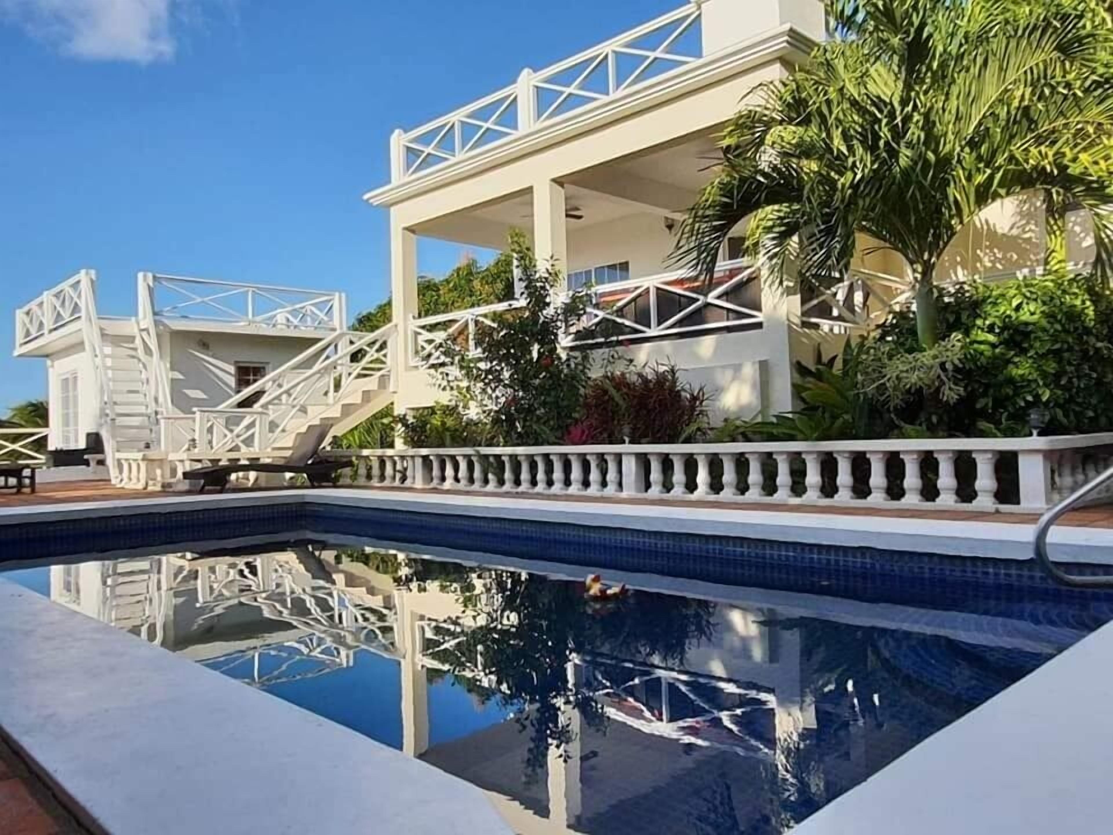 A pool with a white balustrade, a staircase leading to a covered area, and lush greenery.