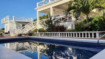 A pool with a white balustrade, a staircase leading to a covered area, and lush greenery.