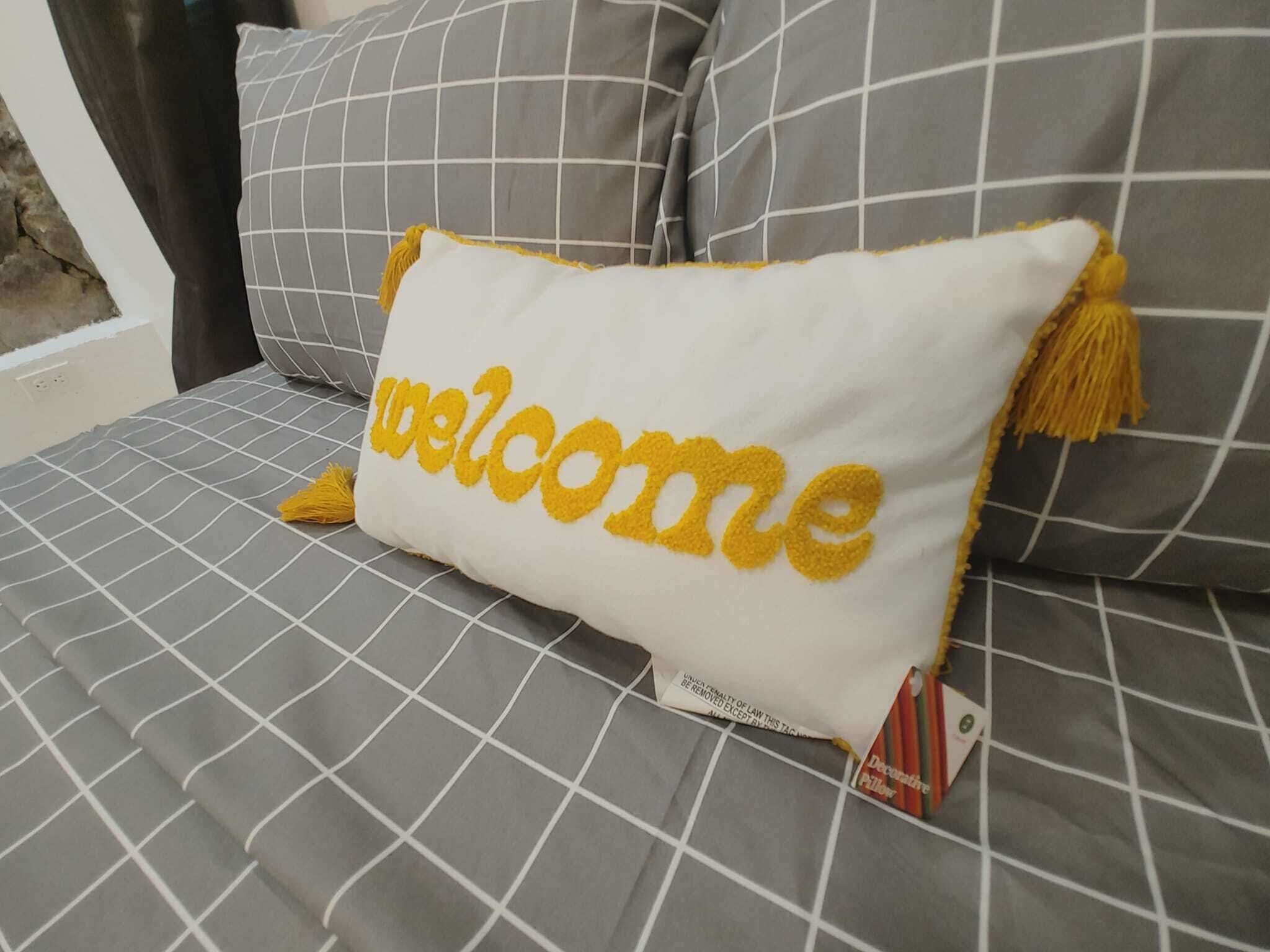 A bed with a white pillow featuring the word "welcome" in yellow.