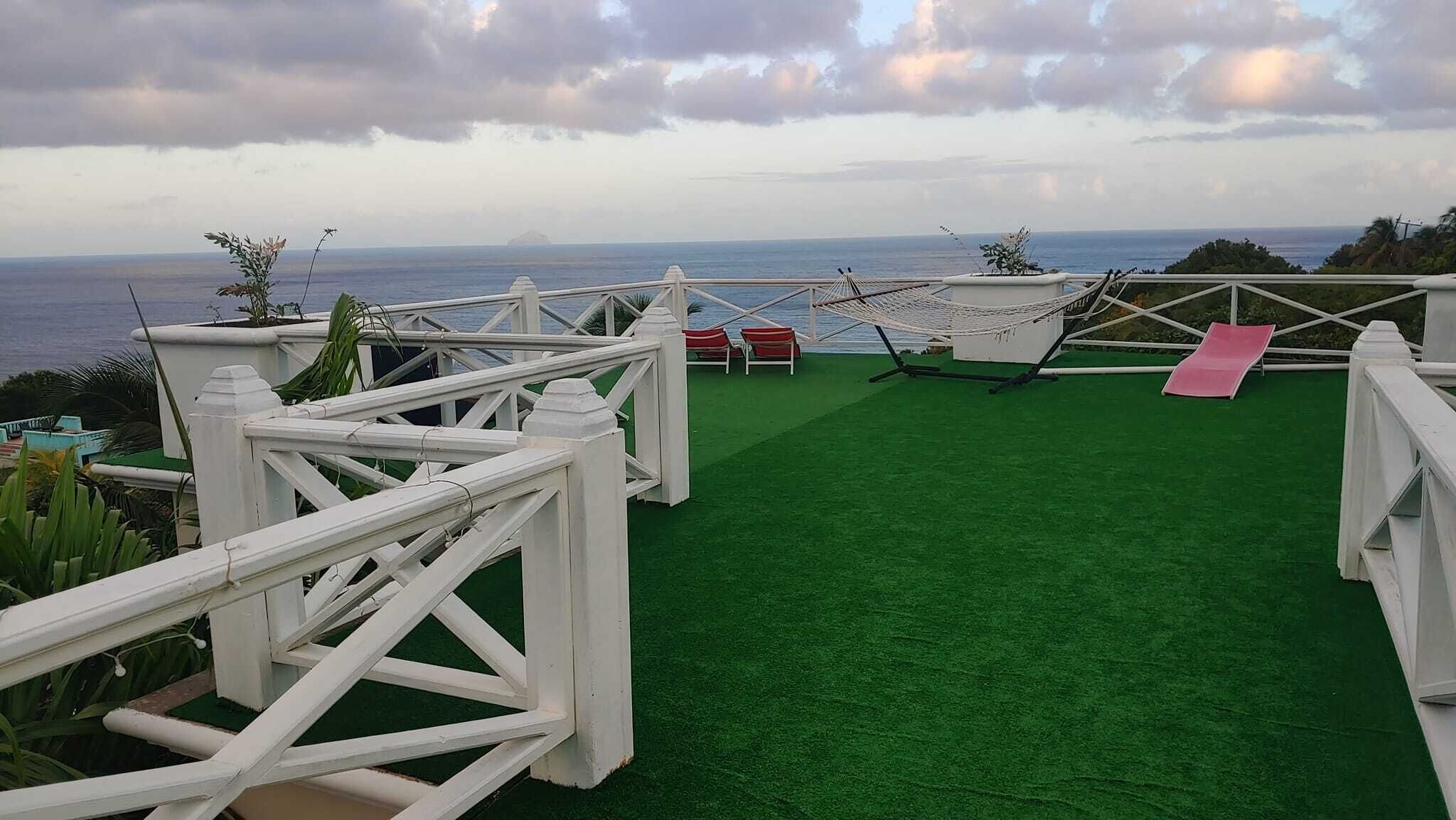 A rooftop terrace with a view of the ocean, green artificial turf, white railings, and a hammock.