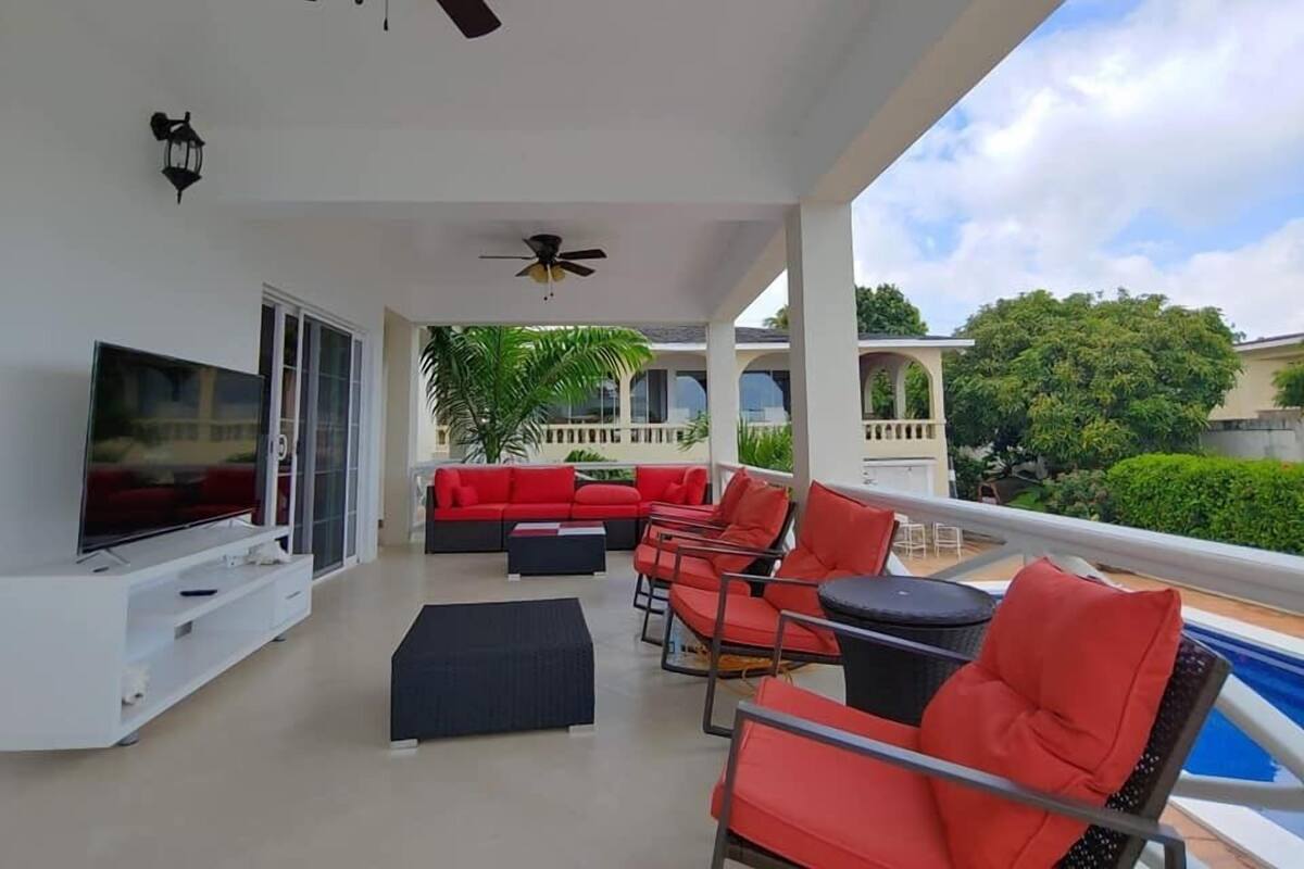 A covered outdoor area with a TV, red and black furniture, ceiling fans, and a pool.