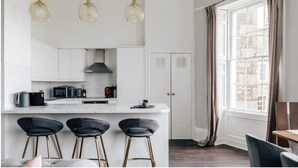 A modern kitchen with white cabinets, a marble countertop, and a central island with bar stools. There is a window with curtains and a view of the city. - The Portland Apartment, 2 beds with parking (Bath)