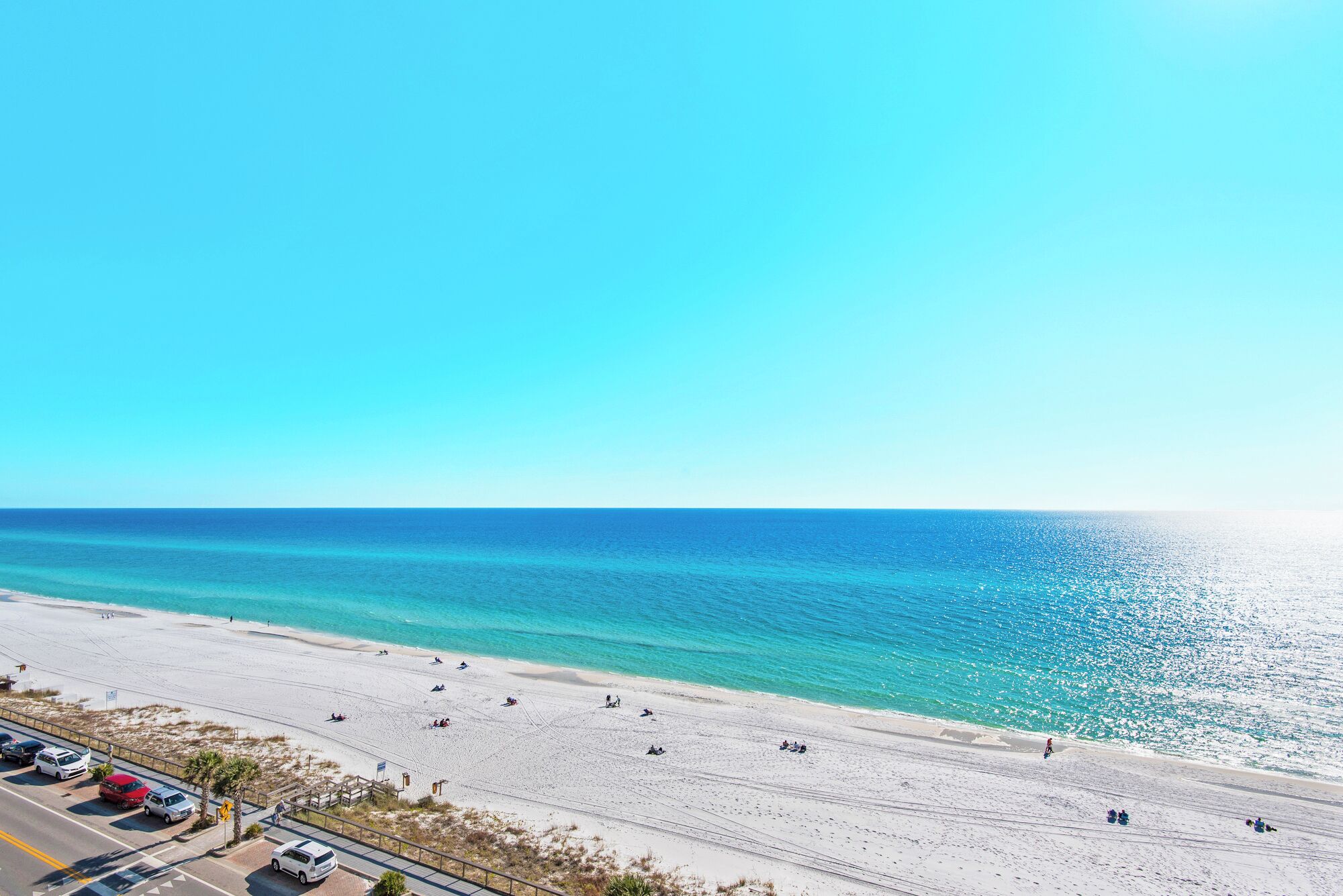 A beach with clear blue water, a sandy shore, and a road with parked cars.
