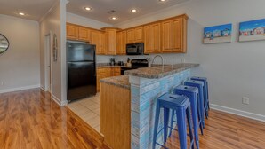A modern kitchen with wooden cabinets, a stainless steel refrigerator, a microwave, and a granite countertop. There are four blue bar stools and a painting of beach houses on the wall. - 4 Bedroom, 3 1/2 Bath Dog-Friendly Beach Home with Private Pool (Garden City Beach)