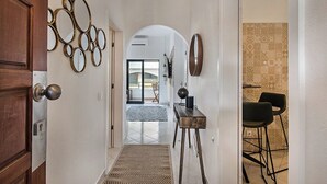 Um corredor com porta de madeira, espelho fixado na parede, pequena mesa com abajur e tapete estampado.
