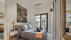 Quarto moderno com uma cama grande, mesa de cabeceira, abajur, obra de arte na parede e vista para o exterior por uma porta de vidro.