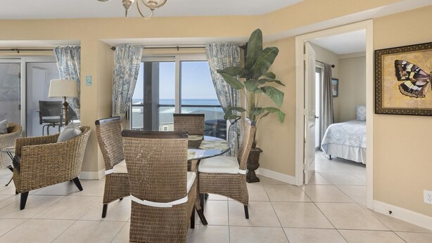 A room with a dining table and chairs, a large potted plant, a bed, and a painting on the wall. - 1 Bedroom, 1 Bath Gulf Front Condo t Regency Towers 603 East! (Pensacola Beach)