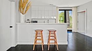 Une cuisine moderne avec des meubles blancs, un îlot central doté de tabourets et une vue sur la verdure à l’extérieur.