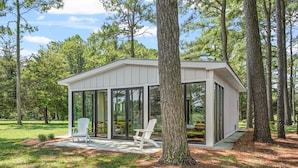 Une cabane moderne dotée de grandes portes vitrées, de deux fauteuils Adirondack blancs et entourée d’une forêt.
