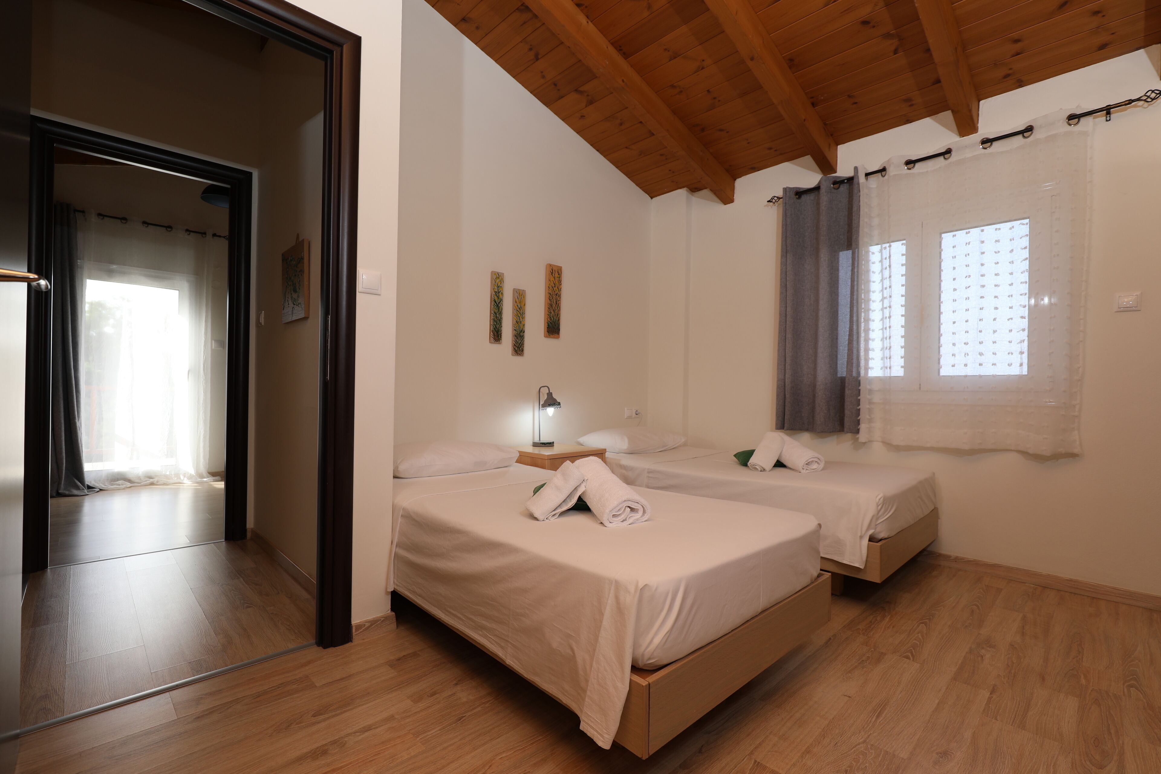 A bedroom with two beds, wooden flooring, a wooden ceiling, and a window with curtains.