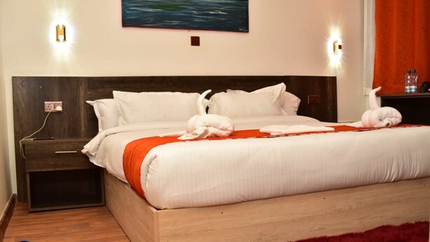 A hotel room with a wooden headboard, white bedding, a red throw blanket, and a red furry rug. There are two wall-mounted lights, a painting, and a water bottle on the nightstand. - Old Station Lounge Naivasha (Naivasha)
