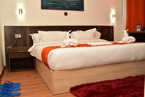 Habitación de hotel con cabecera de madera, ropa de cama blanca, manta roja y alfombra mullida de color rojo. Se observan dos lámparas de pared, un cuadro y una botella de agua sobre la mesita de noche.