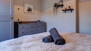 En snyggt bäddad säng med en hoprullad handduk, en grå byrå med lådor, en liten växt på byrån och en vägghylla med en tavla och en växt.