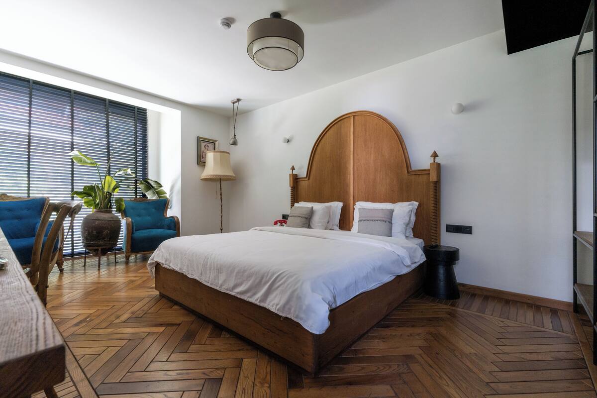 A modern bedroom with a large bed, wooden headboard, and a view of the outdoors through large windows.