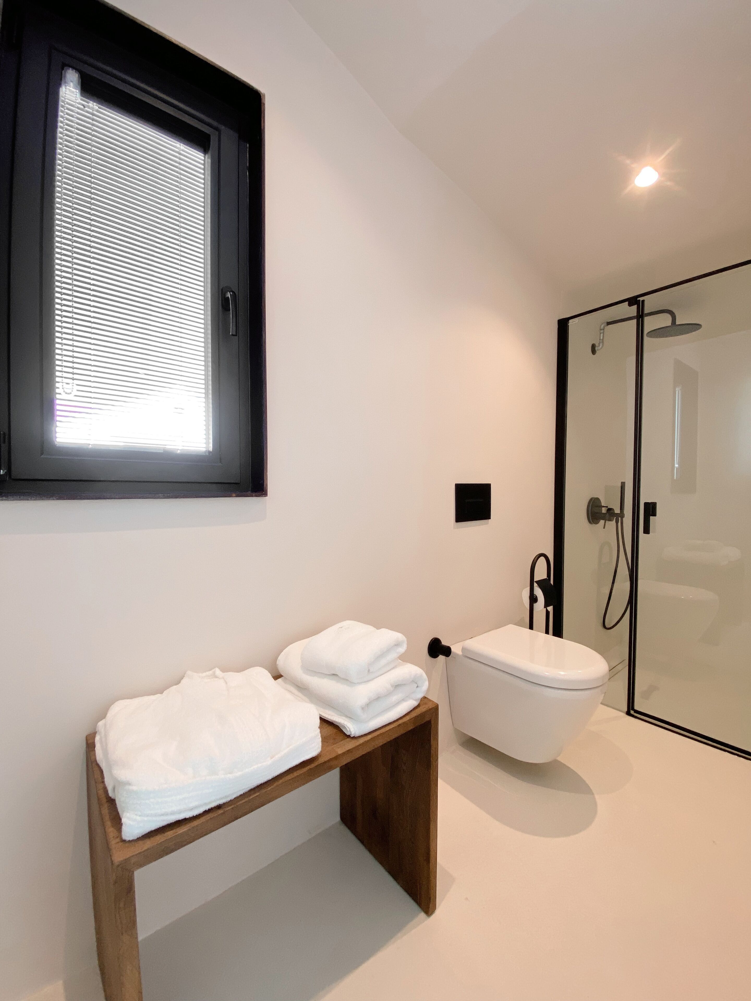 A modern bathroom with a toilet, a glass-enclosed shower, and a wooden bench with folded towels.