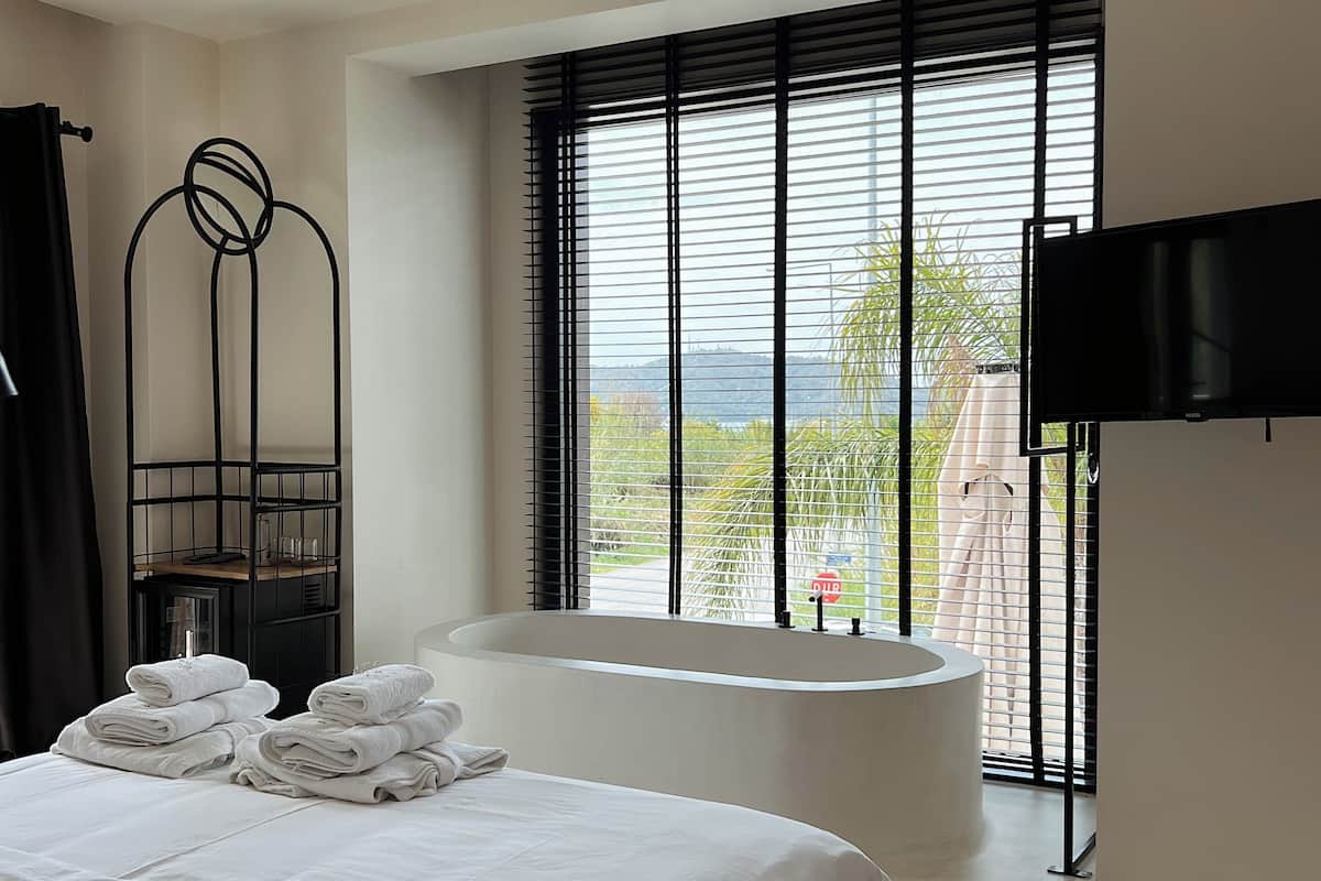 A modern hotel room with a large bed, a bathtub, a TV, and a window with blinds.