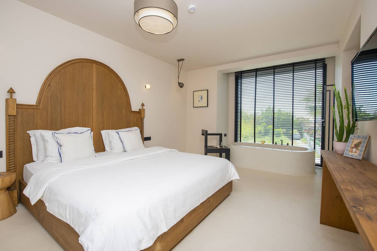 A bedroom with a large bed, a wooden headboard, a window with blinds, a small table, and a potted plant.