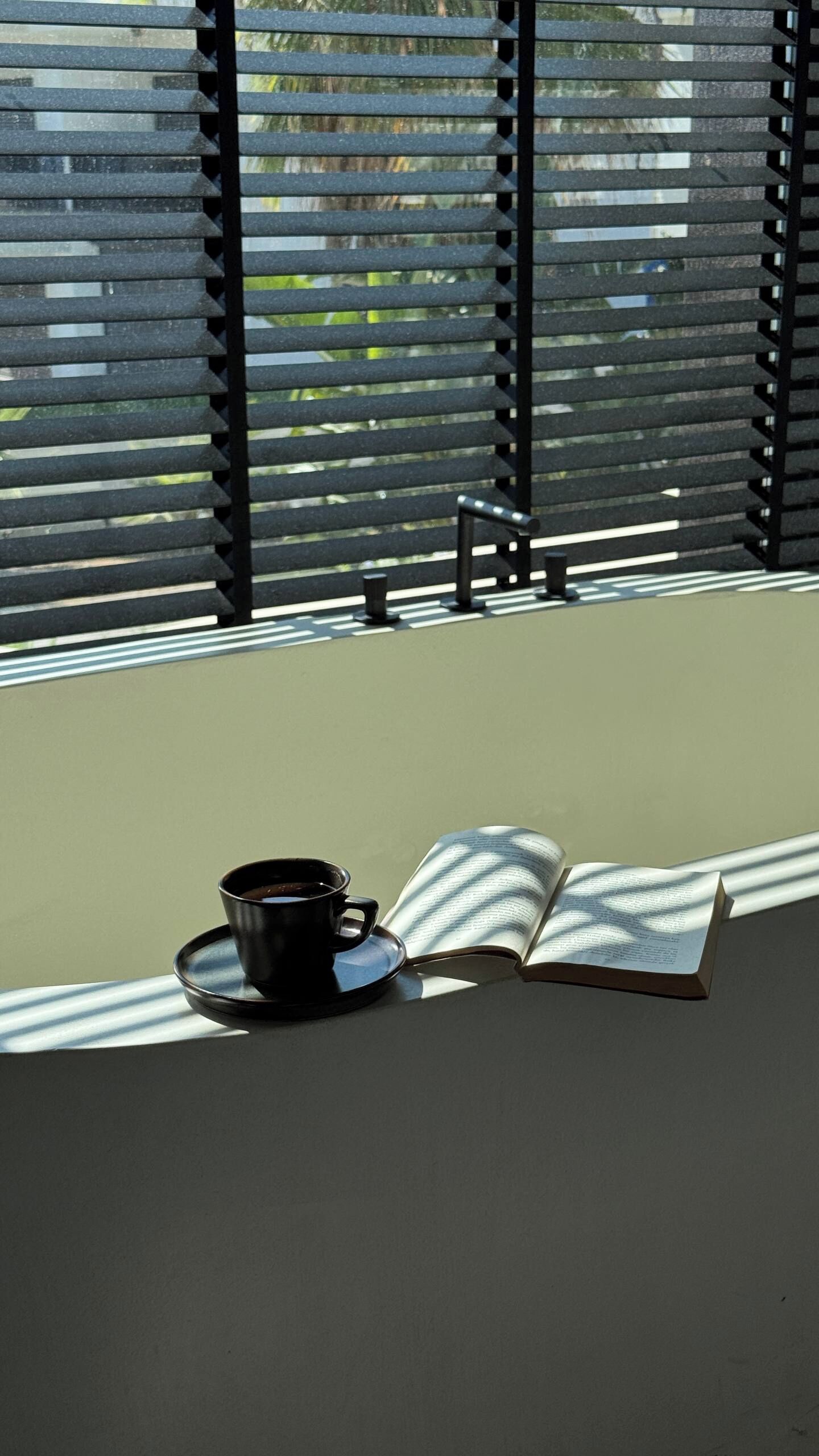 Een raam met horizontale zonwering, een beker en een open boek op een tafel.