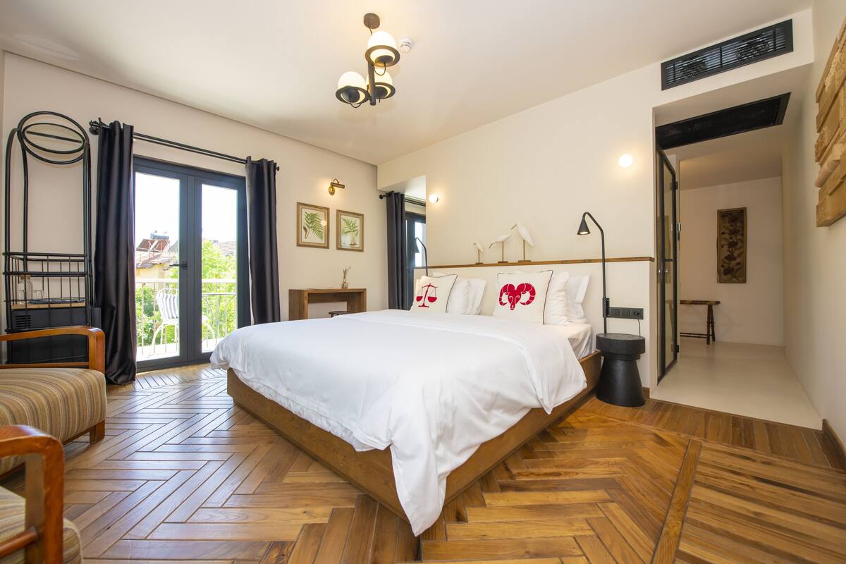 A bedroom with a wooden herringbone floor, a large bed with white bedding, a bedside table, a chair, and a view through a sliding glass door to the outside.