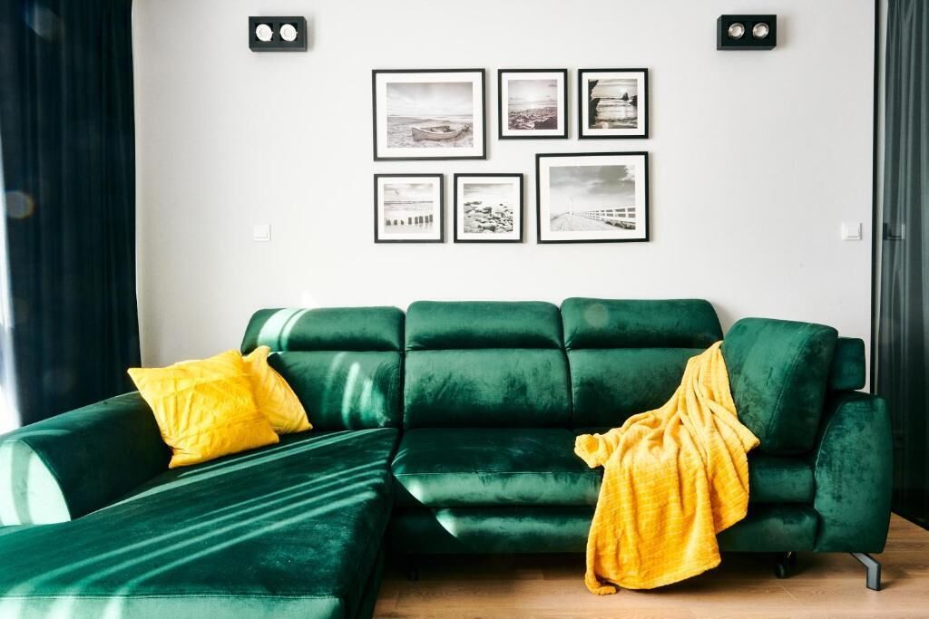 A green sofa with yellow pillows and a yellow blanket, a white wall with framed pictures, and two black wall-mounted lights.