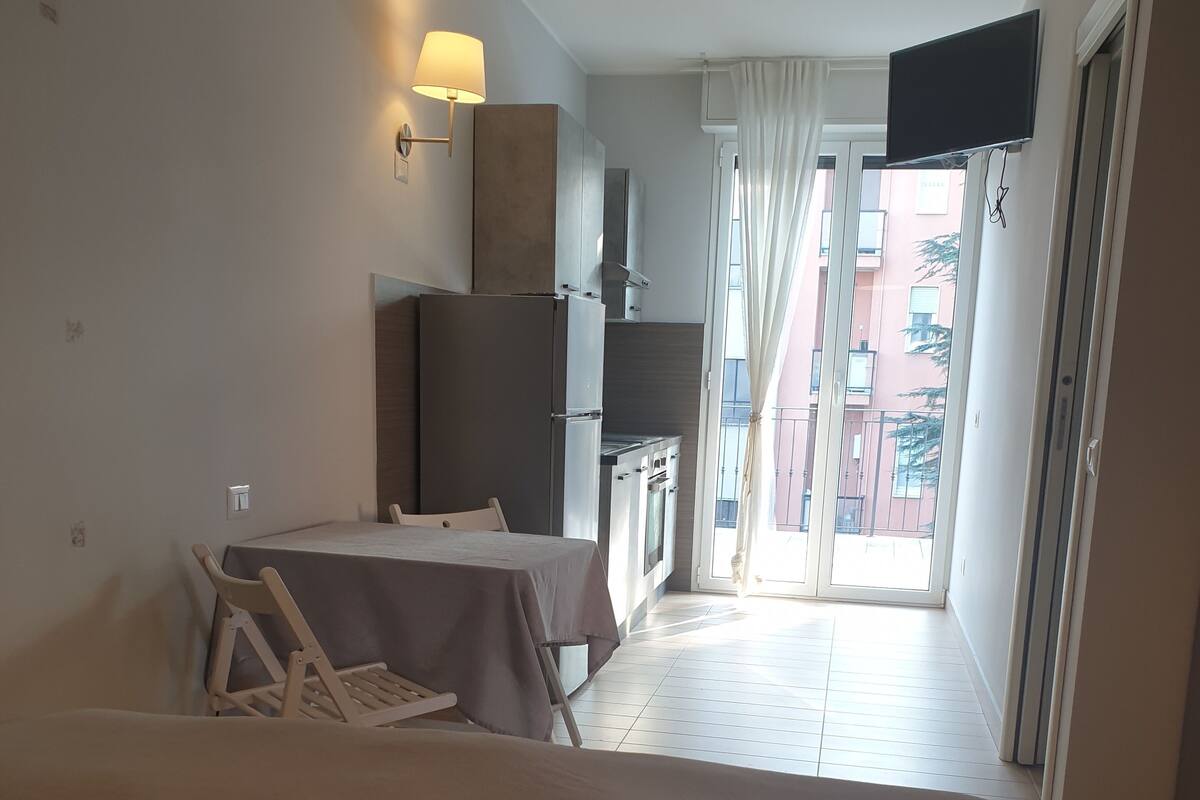 A hotel room with a bed, a table with chairs, a kitchenette, and a balcony with a view of buildings.