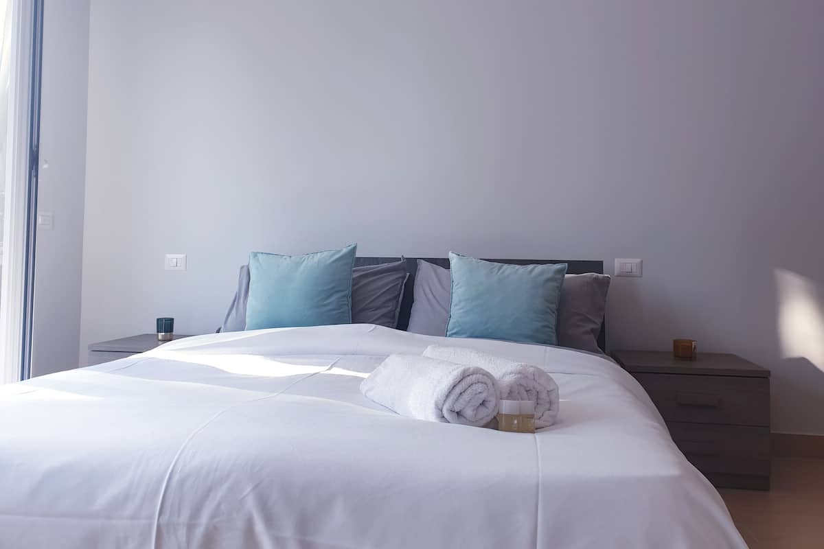 A neatly made bed with white linens and rolled towels, a bedside table with a cup, and a nightstand with a lamp.