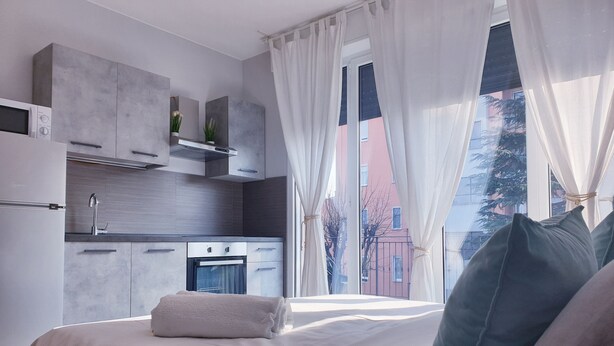 Une chambre moderne avec un lit, des oreillers, une fenêtre avec des rideaux, un coin cuisine avec des placards et des appareils électroménagers, et une vue sur les bâtiments à l’extérieur.