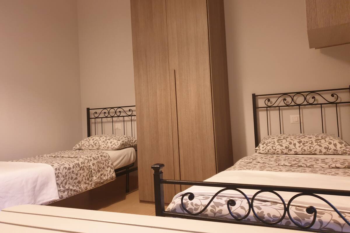 A room with two beds, each with a patterned bedspread and matching pillows. There is a wooden wardrobe and a metal headboard with a decorative design.