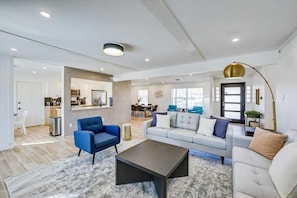 A spacious living area with a sofa, armchairs, and a coffee table. There is a kitchen in the background, a dining area, and a large window providing natural light.