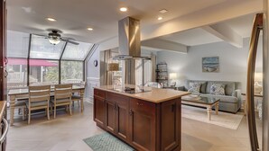 Una cocina amplia con una isla central, un comedor con sillas de madera, una sala de estar con sofá y mesa de centro, y vistas al exterior a través de grandes ventanales.