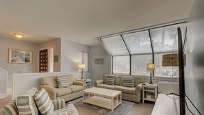 Una sala de estar con un sofá, una mesa de centro y un mueble para televisor. Hay dos lámparas sobre mesitas auxiliares y un ventanal con persianas.