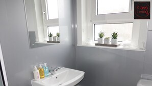 A bathroom with a white sink, mirror, and window. There are toiletries on the sink and a small trash bin.